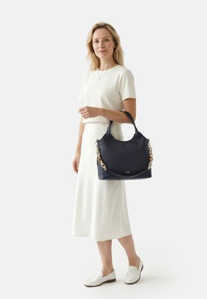 Frau in weißem Kleid und Mokassins, die eine große schwarze Lederhandtasche mit einem groben Kettengurt hält.