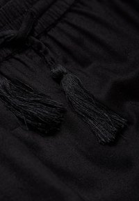 Gros plan sur un tissu noir avec deux pompons texturés au niveau de la taille, présenté avec une finition lisse et mate et un détail froncé.
