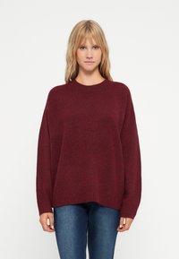 Burgundy oversized tröja med rund hals, ribbad nederkant och mudd, gjord av ett mjukt stickat material, matchad med passande blå jeans.