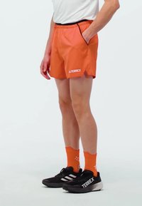 Pantalones cortos atléticos naranjas con cinturilla negra y bolsillos laterales, combinados con calcetines naranjas y zapatillas negras con acentos blancos.