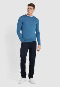 Niebieski sweter robiony na drutach z okrągłym dekoltem, w połączeniu z ciemnymi dżinsami i białymi trampkami. Żebrowane ściągacze i dół podkreślają dopasowanie.