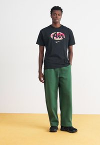Zwarte T-shirt met korte mouwen met een kleurrijk "Air" logo, gecombineerd met losse groene joggingbroek. Model draagt zwarte sportschoenen.