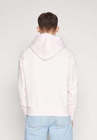 Sudadera rosa claro con capucha amplia, texture suave, puños y dobladillo acanalados, combinada con vaqueros azul claro. Se muestra vista posterior.