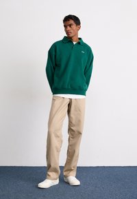 Sweatshirt verde oversized cu guler, purtat peste o cămașă albă. Pereche de pantaloni bej și adidași albi. Detalii minime de branding vizibile.