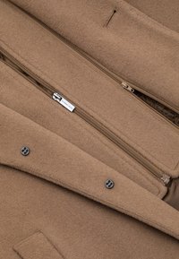 Wnętrze brązowego wełnianego płaszcza z kieszenią na zamek, detalami w postaci guzików i metką. Miękka tekstura z prostym wzorem i jednolitą kolorystyką.