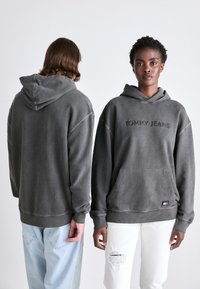 Grå hettegenser med frontlomme, "TOMMY JEANS" preget logo, ribbestrikkede mansjetter og kant. Båret av to modeller, en i lyse jeans, en i hvite jeans.