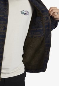 Giacca navale con motivo geometrico marrone, con zip, foderata in pile oliva. T-shirt bianca sotto con logo. Tasca laterale visibile.