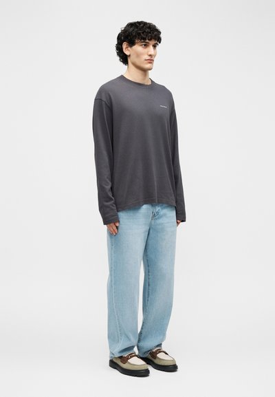 Νέος άνδρας με σγουρά μαλλιά, φοράει μακρυμάνικο πουκάμισο σκούρου γκρι, ανοιχτόχρωμο μπλε φαρδύ τζιν και καφέ-μπεζ παπούτσια, στέκεται αντικρίζοντας προς τα εμπρός.