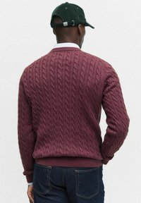 Vinröd kabelstickad tröja med ribbad ärm- och nederkant. Bärs över en vit skjorta med krage, tillsammans med mörka denimjeans. Grön keps på huvudet.