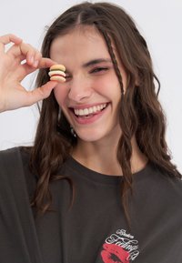 Een glimlachend persoon houdt een kleine, driedelige macaron dessert voor hun oog. Ze dragen een donker t-shirt met een rode lippen graphic.