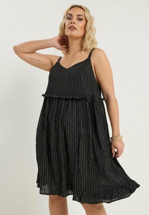 Frau mit blonden Haaren, die ein ärmelloses schwarzes Kleid mit dünnen weißen vertikalen Streifen und Rüschendetails trägt, posiert mit einem erhobenen Arm.