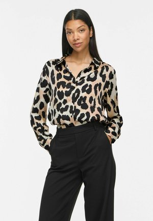 Kvinde med langt mørkt hår iført en beige og sort leopardmønstret bluse, stoppet ned i sorte højtaljede bukser med hænderne i lommerne.