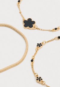 Zlaté náramky s čiernymi smaltovými kvetinovými príveskami a hladkým zlatým hadím reťazcom na bielom pozadí.