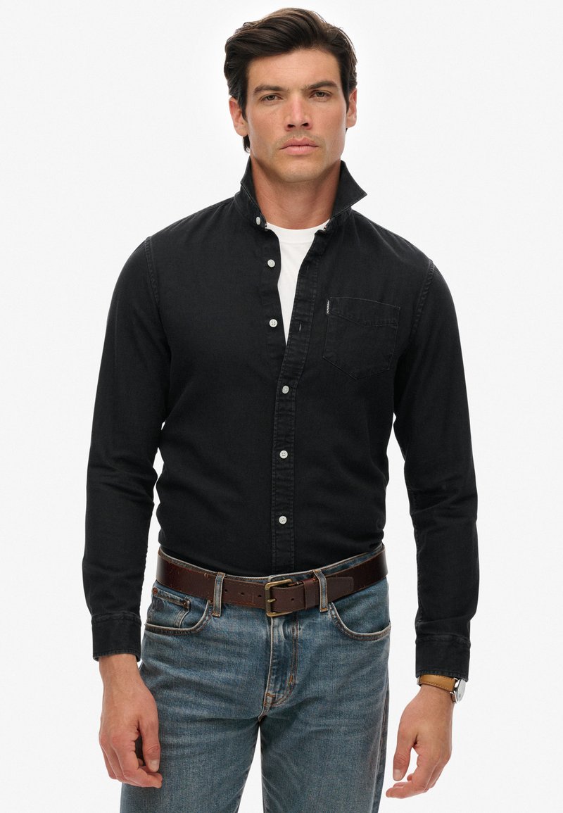 Homme portant une chemise noire boutonnée avec col relevé, t-shirt blanc en dessous, jean bleu, ceinture en cuir marron et montre-bracelet, debout face à l'avant.