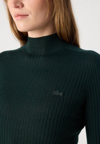 Pull col roulé côtelé en vert foncé, en tissu doux avec une coupe ajustée. Doté d'un logo ton sur ton discret sur la poitrine.