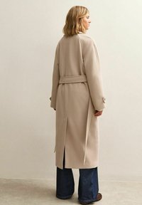 Długi płaszcz w jasnobeżowym kolorze z paskiem w talii, opuszczanymi ramionami, rozcięciem z tyłu oraz dużymi, zaokrąglonymi guzikami na rękawach. Noszony na szerokich, niebieskich dżinsach.