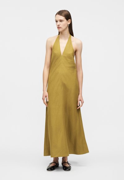 Γυναίκα που φοράει μακρύ, ελαιόχρωμο φόρεμα χωρίς μανίκια με λαιμόκοψη halter, και μαύρες μπαλαρίνες, στέκεται μπροστά σε ένα απλό φωτεινό φόντο.