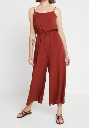 Combinaison rouge sans manches avec une coupe décontractée, taille élastique, jambes larges et tissu texturé. Accentuée par de fines bretelles. Design décontracté.