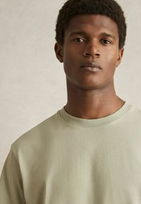 Tricou din bumbac de culoare verde deschis, cu guler rotund și mâneci scurte. Textură fină, design minimal, fără modele sau grafice vizibile.