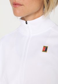 Gros plan d'une personne portant une veste blanche zippée avec un petit logo coloré sur la poitrine gauche et des lèvres brillantes visibles.
