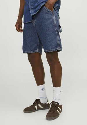 Pantalones cortos de mezclilla en azul oscuro con dobladillo deshilachado, bolsillos laterales y pasadores para cinturón, combinados con zapatillas de gamuza marrón y calcetines blancos con un logo de cara sonriente.