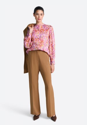 Woman wearing a pink floral blouse, brown high-waist trousers, holding a matching brown jacket over her shoulder, standing on white background.