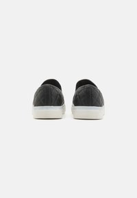 Chaussures noires sans lacets matelassées avec un bout arrondi et une semelle en caoutchouc blanche. Tige texturée avec une finition lisse ; sans logos ni pièces métalliques visibles.