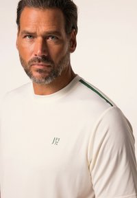Camisa de manga corta color crema en tejido suave, con un logo verde en el pecho y rayas verdes en los hombros.