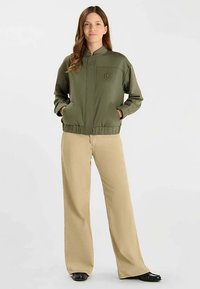 Veste bomber vert olive avec col montant, fermeture éclair avant, poches latérales et bordure élastique ; associée à un pantalon large crème.