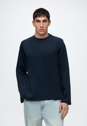 Jeune homme aux cheveux courts et foncés portant une chemise à manches longues bleu marine et un jean bleu clair, debout devant un fond blanc uni.