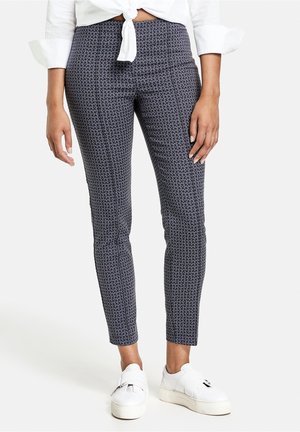 Femme portant un pantalon bleu marine ajusté à motifs, une chemise blanche nouée et des baskets compensées blanches sur fond blanc.