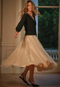 Schwarzer funkelnder Pullover mit langen Ärmeln, cremefarbener plissierter Satinenrock und schwarze Riemchen-Flats. Das Model dreht sich in einem gut beleuchteten Raum mit Holzböden.