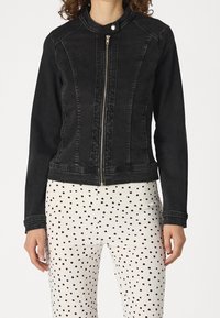 Žena v těsně padnoucí černé bundě na zip s klopou a bílých kalhotách s nepravidelnými černými puntíky.