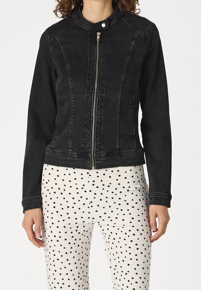 Žena v těsně padnoucí černé bundě na zip s klopou a bílých kalhotách s nepravidelnými černými puntíky.