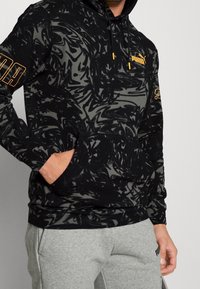 Czarna i szara bluza z kapturem o wirującym wzorze, z kieszenią kangurką, sznurkiem przy kapturze oraz żółtym logo Pumy na klatce piersiowej i rękawie.