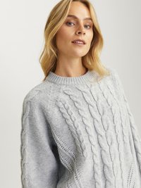 Svetlo siva pletena jopica s krojem okoli vratu, rebrastimi manšeti in teksturiranim dizajnom z opletenimi vzorci na rokavih. Mehki material.