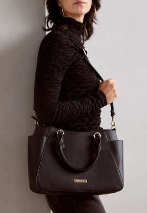 Woman wearing a textured black long-sleeve top holding a structured black handbag with gold hardware and a shoulder strap.