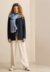 Giacca in pile blu navy con due bottoni, sciarpa azzurra chiara drappeggiata intorno al collo, pantaloni a gamba larga crema e scarpe da ginnastica chiare con suola texture.
