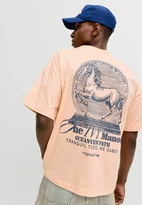 Persoană purtând un tricou piersică cu o grafică a unui cal ridicat pe picioarele din spate, de culoare bleumarin, și text pe spate, asortat cu pantaloni scurți din denim deschis și o șapcă albastră.