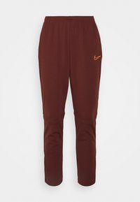 Bruine Nike-sweatpants met een elastische tailleband, toelopende benen en een oranje logo-detail op de linkerdij. Zachte stoftextuur.