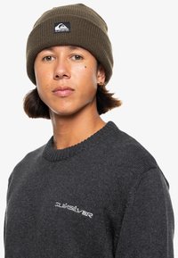 Mörkgrå stickad tröja med ribbad rund hals och broderad logotyp. Oljgrön mössa med en märkeslapp på framsidan.