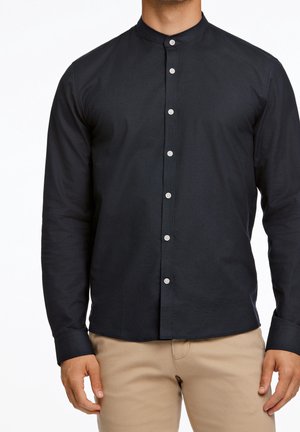 Mężczyzna w czarnej koszuli z długim rękawem i mandaryńskim kołnierzykiem zapinanej na guziki oraz beżowych spodniach, stojący na białym tle.