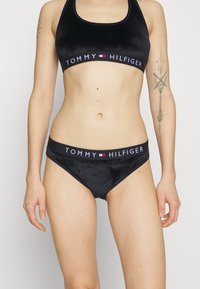 Svart velour sportbh och matchande trosor med en märkeselastisk band som har "TOMMY HILFIGER" i vit text och färgglada detaljer.