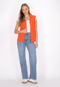 Orange ärmellose Zip-Weste über einem weißen Hemd, kombiniert mit hellblauen weit geschnittenen Jeans und weißen Sneakers. Einfaches Design mit modernem Schnitt.