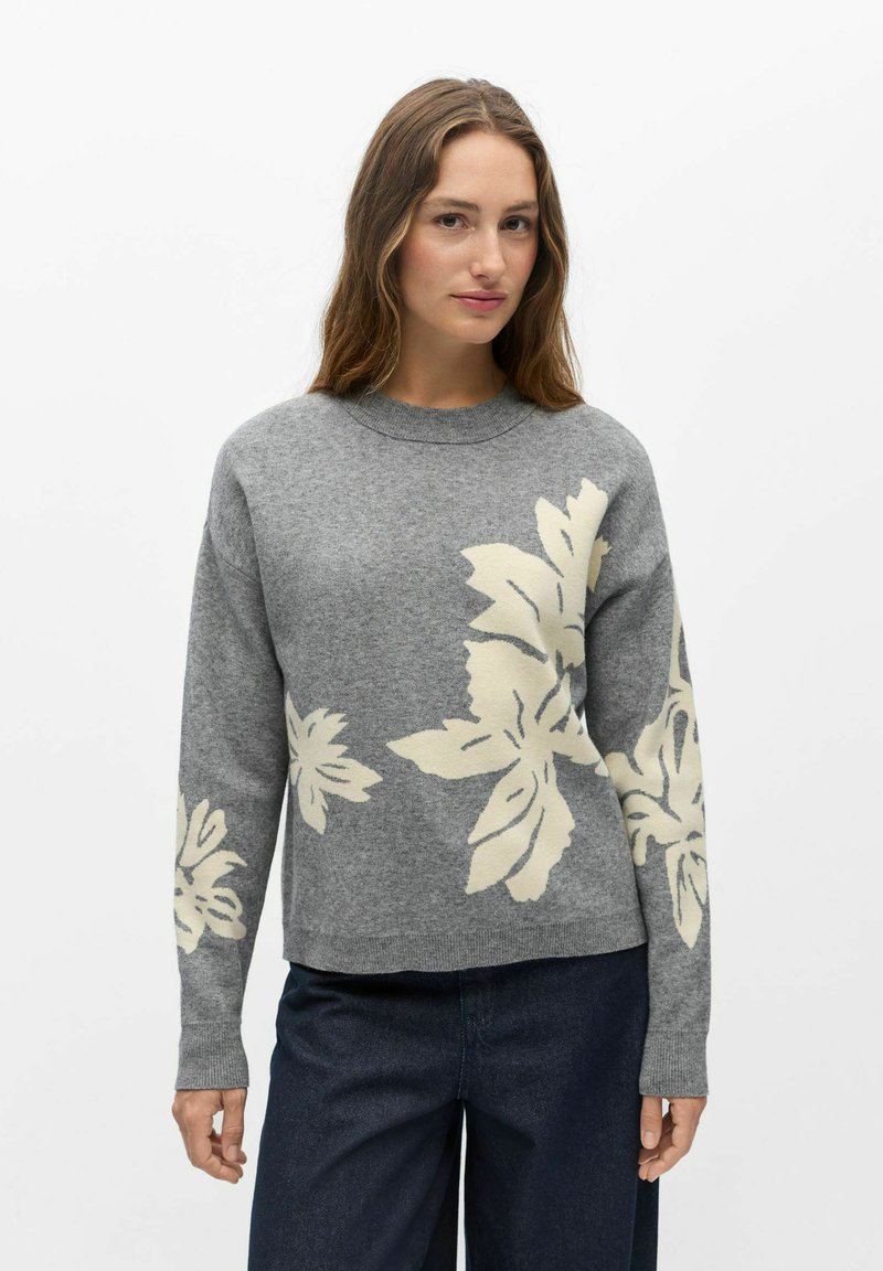 Femme aux longs cheveux bruns portant un pull gris avec de grands motifs floraux blancs et un pantalon bleu foncé sur un fond blanc uni.