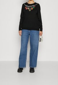 Black knit sweater with white trim and embroidered holiday ornaments on the neckline, paired with light blue wide-leg jeans and black shoes.