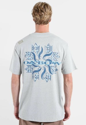 Homme aux cheveux blonds courts portant un T-shirt gris clair avec un grand logo bleu Oxbow au dos.