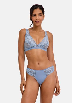 Frau trägt hellblaues Spitzen-Dessous-Set mit Blumenmustern, steht mit entspannter Haltung vor einem schlichten hellen Hintergrund.