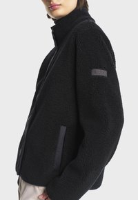 Μαύρο φλις μπουφάν με υφασμάτινη επιφάνεια, ψηλό γιακά και ένα λογότυπο στην αριστερή μανσέτα. Διαθέτει πλευρικές τσέπες και χαλαρή εφαρμογή.