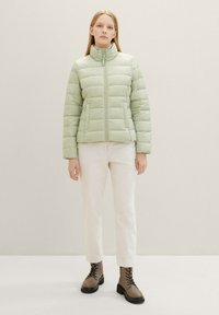 Hellegrüne Daunenjacke mit horizontalem Quiltmuster, hohem Kragen und Frontreißverschluss, kombiniert mit weißen Hosen und braunen Schnürstiefeln.